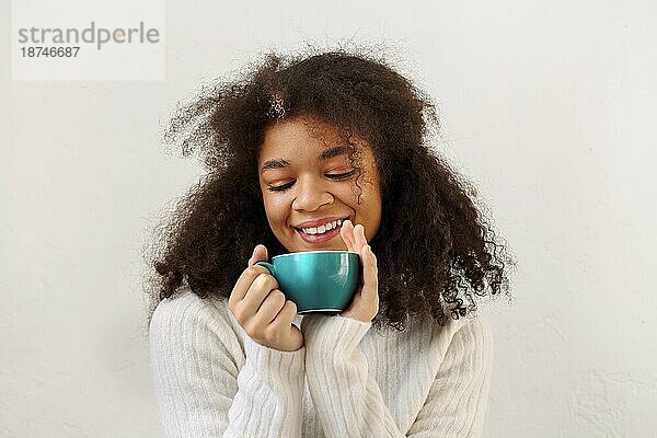 Glückliches  erfreutes  lockiges afrikanisches amerikanisches Mädchen mit geschlossenen Augen  das eine Tasse Tee genießt und sich in der kalten Wintersaison entspannt  mit einem warmen  gestrickten weißen Pullover  vor weißem Hintergrundem Studiohintergrund