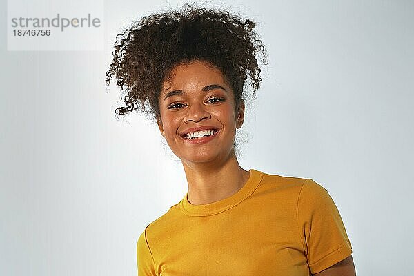 Überglückliche afroamerikanische Frau in leuchtend gelbem TShirt mit lockigem Haar  die lachend in die Kamera schaut und ihre weißen  glatten Zähne zeigt  posiert vor einem grauen Studiowandhintergrund. Glück Konzept
