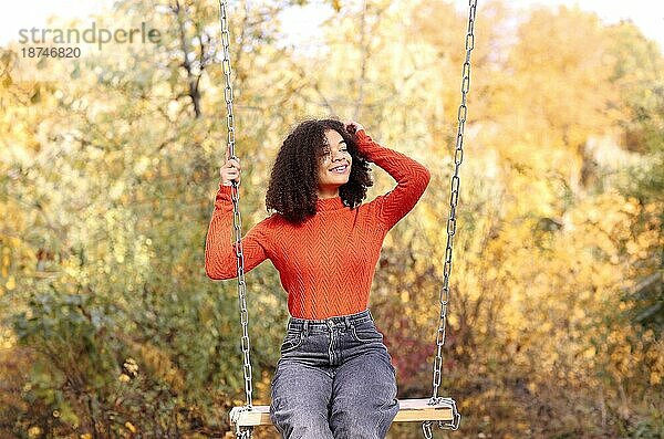 Einheit mit der Natur. Junge glückliche sorglose afrikanische amerikanische Frau mit lockigem Haar in orange warmen Pullover sitzen auf Schaukel in schönen goldenen Herbst Wald