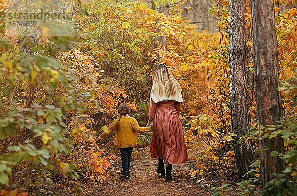 Rückansicht einer jungen Familie  Mutter und kleine Tochter  auf einem Spaziergang im Herbstwald  Mutter und Kind halten sich an den Händen und gehen einen mit gelben Blättern bedeckten Weg entlang  sie verbringen Zeit in der schönen Natur