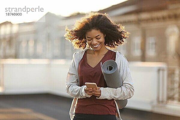 Junge  sportliche  fröhliche afroamerikanische Frau in Sportkleidung mit einer Yogamatte in den Händen  die auf dem Dach steht  ihr Handy benutzt und lächelt  sie unterhält sich mit Freunden vor dem morgendlichen Sporttraining auf dem Dach