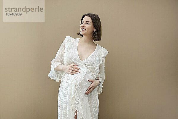 Attraktive schwangere Frau im weißen Kleid auf beigem Hintergrund. Mutterschaft und Gesundheitswesen Konzept