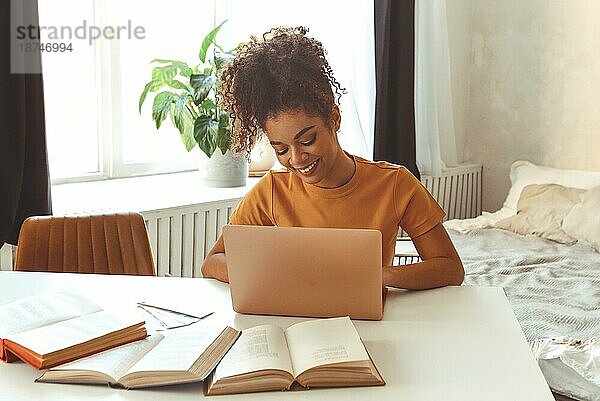 Fröhliche junge afroamerikanische Studentin  die zu Hause am Schreibtisch sitzt  umgeben von Lehrbüchern  vor einem Laptop  der fleißig studiert und sich auf Prüfungen im Internet vorbereitet  und mit einem zufriedenen Lächeln in den Laptop schaut
