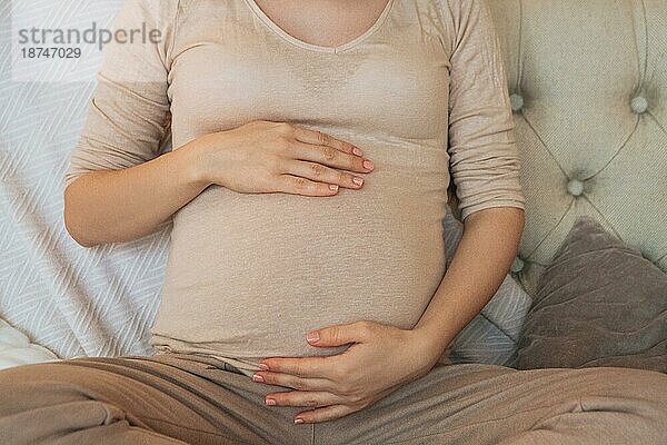 Cropped Schuss der schwangeren Frau hält Bauch beim Entspannen auf der Couch zu Hause  werdende Mutter sitzt auf dem Sofa in Innenräumen und sanft berühren Bauch. Schwangerschaft  Mutterschaft und pränatale Periode Konzept