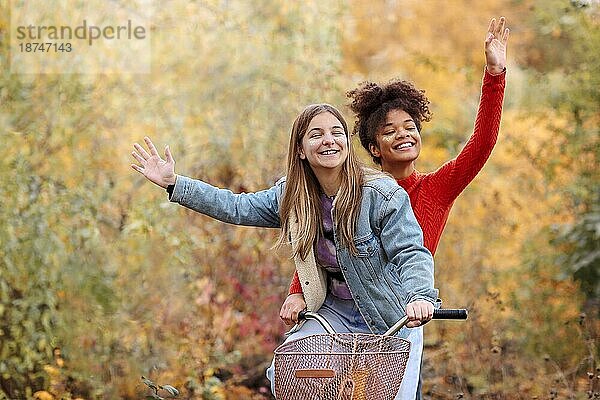 Zwei junge glückliche verschiedene weibliche beste Freunde fahren Fahrrad zusammen im Herbst Wald  aktive positive multirassische weibliche Freunde lächelnd in die Kamera und wavinh Hände beim Fahren Stadt Fahrrad im Herbst Natur