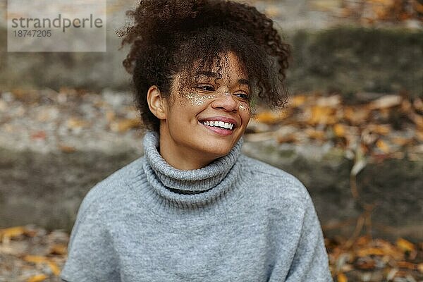 Porträt einer jungen  schönen  glücklichen afroamerikanischen Frau mit glamourösem  glänzendem Make up und in einem warmen Pullover  die bei bewölktem Herbstwetter im Freien sitzt und breit und fröhlich lächelnd in die Ferne schaut