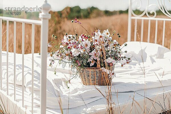 Viele schöne bunte Wildblumen in Weidenkorb auf dem Bett mit sauberen weißen Bettwäsche in Getreide Weizen Sommerfeld oder Wiese auf sonnigen Sommertag  selektiver Fokus