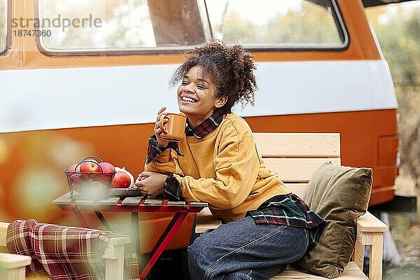 Junge afroamerikanische Frau sitzt am Tisch in der Nähe von Auto im Herbst Wald und trinken heißen Tee  verträumt gemischter Rasse weiblichen entspannenden rund um Campingplatz  Zeit in der Natur. Camping  Reisen und Vanlife