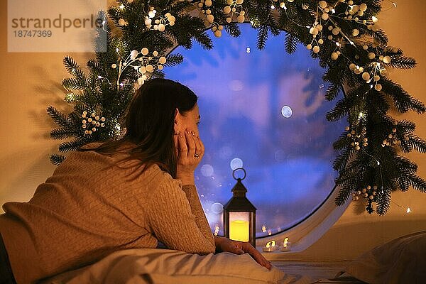 Weihnachtswunder. Rückansicht einer verträumten Frau  die aus einem runden Fenster schaut  um einen Weihnachtsstern in der Nacht zu sehen  eine Frau  die sich auf der Fensterbank mit einer beleuchteten Laterne entspannt  gemütliche magische Atmosphäre zu Hause in der Weihnachtszeit