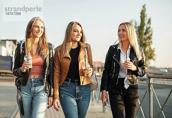 Glückliche junge Frauen mit Getränken zum Mitnehmen  die auf dem Bürgersteig spazieren gehen und an einem sonnigen Wochenendtag in der Stadt lächeln