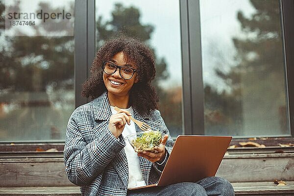 Glückliche afroamerikanische Frau  die Salat ißt und gesund zu Mittag ißt  während sie sich im Freien mit einem Laptop ausruht. Positives schwarzes Mädchen Studentin mit Pause beim Lernen online im Park auf Herbst Tag