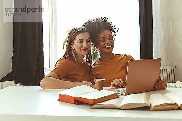 Zwei glückliche Freundinnen  die sich auf Prüfungen vorbereiten  sitzen zusammen vor einem Laptop neben aufgeschlagenen Lehrbüchern vor dem Hintergrund eines Wohnzimmerinterieurs  fröhliche Klassenkameraden  Studenten  die gemeinsam im Haus lernen