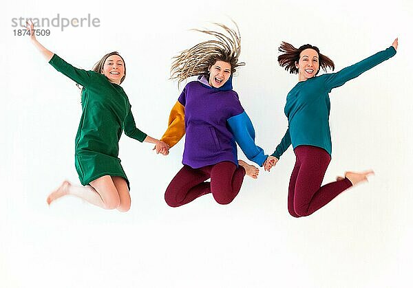 Bild von drei glücklichen verspielten barfüßigen Frauen unterschiedlichen Alters  die sich an den Händen halten  springen und Spaß haben  Urlaub genießen  Leistung feiern  vor weißem Hintergrundem Studiohintergrund