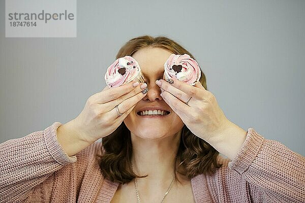 Erfreute weibliche Abdeckung Augen mit leckeren Cupcakes und lächelnd  während Sie Spaß gegen grauen Hintergrund