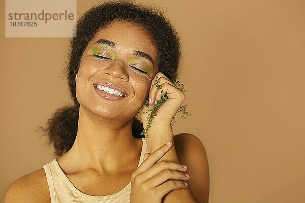 Porträt einer schönen jungen afrikanischen Frau  die mit geschlossenen Augen vor der Kamera posiert und ein hellgrünes Öko Make up zeigt  während sie einen Zweig mit Blättern in der Faust hält und breit lächelt. Öko Bio Kosmetik Konzept