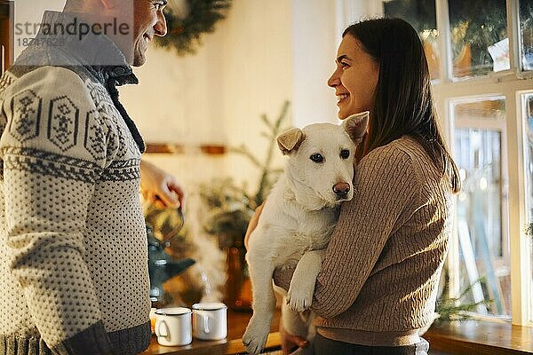 Junges glückliches Paar  Mann und Frau  die einen süßen weißen Hund halten und sich umarmen  während sie in der weihnachtlich geschmückten Küche stehen  schöne Familie mit Welpe  die die Winterferien zusammen in einem gemütlichen warmen Haus genießen