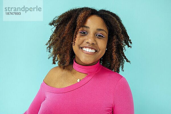 Verträumte junge schöne afroamerikanische Frau mit hellen Augenlinien  die einen rosafarbenen Body über einer blaün Wand trägt  schaut mit glücklichem Gesichtsausdruck  hat ein zahniges Lächeln