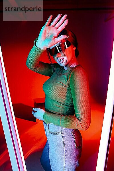 Junge Frau mit futuristischer Brille gestikuliert und schützt ihr Gesicht vor hellem Neonlicht in einem modernen Raum
