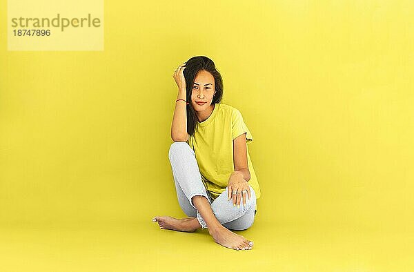 Ganzkörperdarstellung einer asiatischen Frau in Freizeithemd und Jeans  die barfuß auf dem Boden in einem hell erleuchteten gelben Studio sitzt