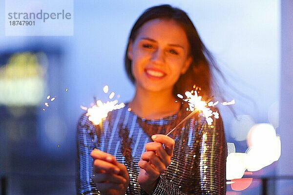 Entzückte junge Dame im Abendkleid  die lächelt und mit brennenden Wunderkerzen während einer nächtlichen Weihnachtsfeier winkt