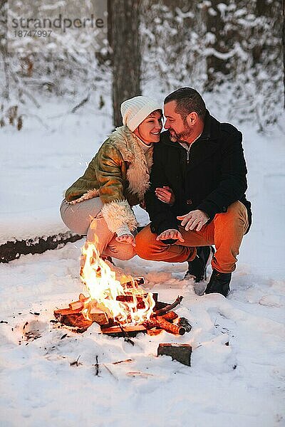 Joyful glückliche Familie Paar in der Liebe Sonnenbaden am Lagerfeuer draußen im Winter verschneiten Wald  Mann und Frau wärmen sich umarmen und liebevollen Blick auf einander. Trekking  Abenteuer und saisonale Urlaub