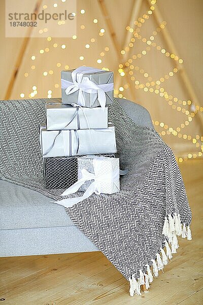 Stapel stilvoll verpackter Geschenkkartons auf der Couch mit Decke in einem gemütlichen Raum an der Wand mit Lichterketten am ersten Weihnachtstag