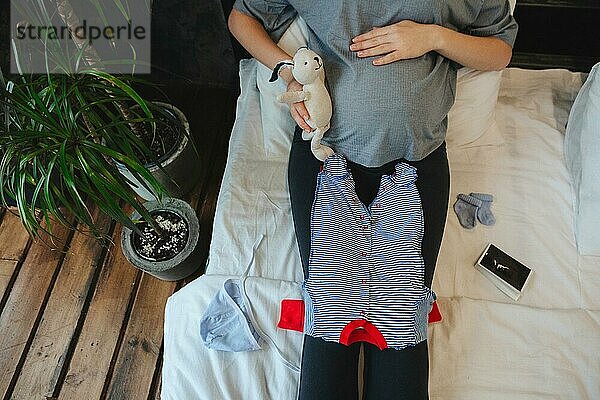 Cropped Schuss der schwangeren Frau Blick auf niedliche Babykleidung während auf dem Bett zu Hause ruht  weibliche werdendes Kind genießen Schwangerschaft  die Vorbereitung für die Elternschaft und der Geburt. Adorable neugeborenen Kleidung