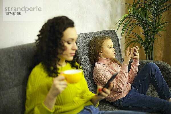 Fokussierte junge Mutter und ihre kleine Tochter benutzen Mobiltelefone  während sie getrennt auf dem Sofa zu Hause sitzen  selektiver Fokus auf das Kind  das sich auf der Couch mit dem Gerät in den Händen entspannt. Technologien im Familienleben