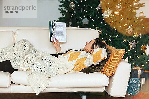 Entspannte Frau mittleren Alters  die sich auf dem Sofa ausruht und ein Buch liest  die sich zu Hause im Wohnzimmer mit geschmücktem Weihnachtsbaum ausruht  eine Frau  die auf der Couch liegt und ihre Freizeit während der Winterferien verbringt