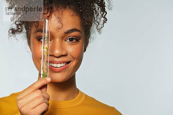 Lächelnde afroamerikanische Frau  die ein Glasreagenzglas mit Microgreens vor dem Gesicht hält  während sie vorgrauem Hintergrund posiert  Platz für Text. Superfood und Ernährung Konzept
