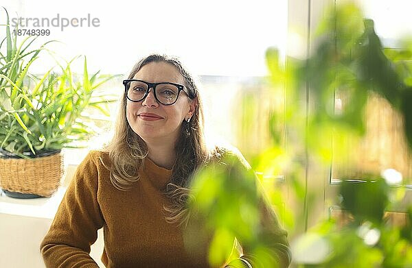 Porträt einer lächelnden Frau mittleren Alters mit Brille  die in einem Raum mit grünen Zimmerpflanzen sitzt  selektiver Fokus. Positive glückliche 40er Jahre Lehrerin mit Brille im Grünen  arbeitet in der Schule