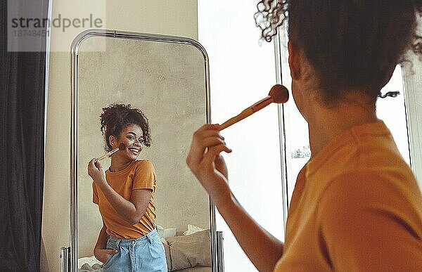 Fröhliche  fröhliche afroamerikanische junge Frau  die sich mit einem Pinsel schminkt  vor einem großen Spiegel zu Hause steht und ihr eigenes Spiegelbild mit einem breiten Lächeln betrachtet. Schönheit Routine Konzept