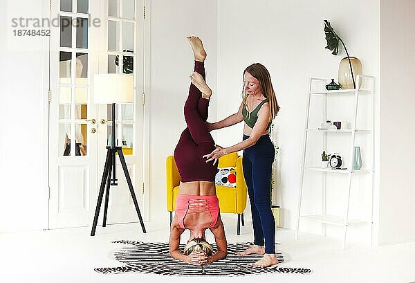 Vollkörper erwachsene Frau unterstützt Freund tun Kopfstand mit Eagle Legs während Yoga Sitzung in gemütlichen Raum zu Hause