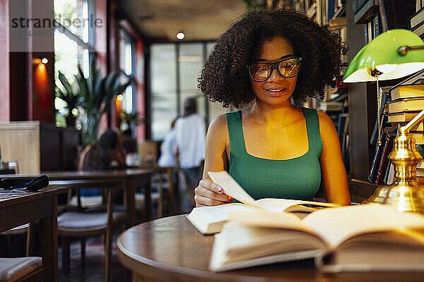 Glückliche afroamerikanische Studentin  die in der Universitätsbibliothek studiert und auf das Buch schaut  mit Brille bei grüner Lampe. Lächelndes Teenagermädchen in gemütlicher Bibliothek