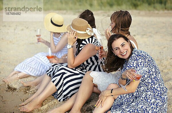 Glückliche Freundinnen in Sommerkleidern  die lächelnd und mit einem Glas Wein anstoßen  während sie sich gemeinsam am Strand ausruhen