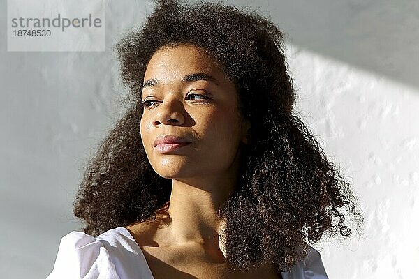 Zartes Porträt einer jungen afroamerikanischen Frau mit lockiger Frisur  die im Sonnenlicht drinnen vor einer grauen Wand steht und mit einem sanften Lächeln zur Seite schaut  Sonnenstrahlen fallen auf das Gesicht einer lächelnden schwarzen Frau