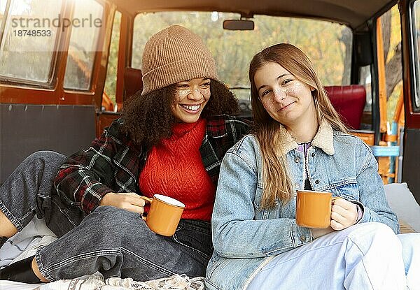 Zwei glückliche multirassische Freundinnen auf dem Kofferraum eines Autos im herbstlichen Wald  die sich umarmen und heißen Tee während einer Abenteuerreise in der herbstlichen Natur trinken  lächelnde Freundinnen  die das Leben genießen  während sie zusammen reisen