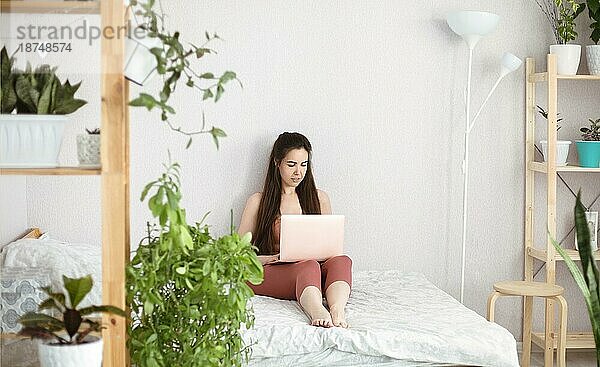 Junge Frau Studentin in Freizeitkleidung mit Laptopcomputer beim Entspannen im Bett im Zimmer mit vielen grünen Pflanzen dekoriert. Fernarbeit von zu Hause aus  Internetshopping oder Online Studium