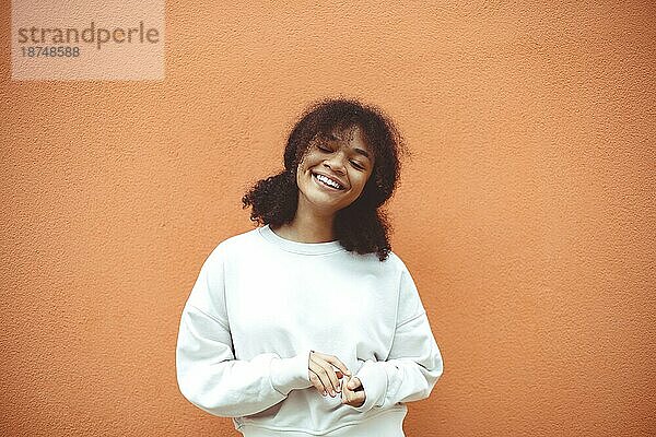 Pures Glück. Nette glückliche afroamerikanische Mädchen mit lockigen Frisur trägt weißen Pullover posiert mit geschlossenen Augen gegen orange Wand. Lächelnde schwarze junge Frau  die mit ihrem Leben zufrieden ist