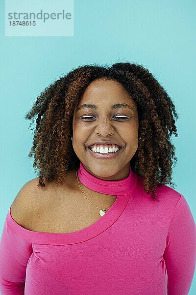 Verträumte junge schöne afroamerikanische Frau mit hellen Augenlinien  die einen rosafarbenen Body über einer blaün Wand trägt  hält ihre Augen geschlossen  schaut mit glücklichem Gesichtsausdruck  hat ein zahniges Lächeln