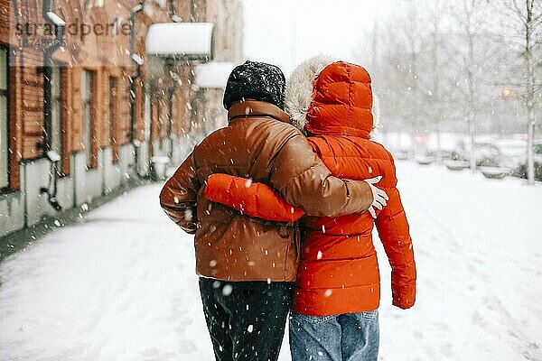 Rückenansicht von anonymen Frauen in Oberbekleidung  die sich gegenseitig umarmen und auf einer verschneiten Straße im Winter laufen