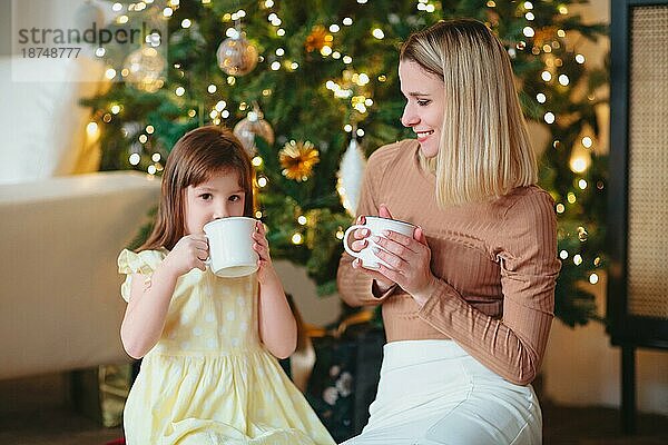 Glückliche Familie  Mutter und kleine Tochter trinken heißen Kakao  sitzen neben dem geschmückten Weihnachtsbaum  Mutter und Kind verbringen Zeit zusammen zu Hause während der Winterferien und feiern das neue Jahr
