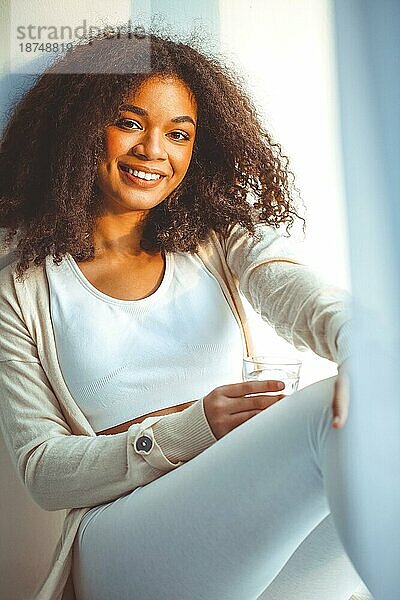 Junge positive afroamerikanische Frau  die mit einem Glas reinem Mineralwasser auf der Fensterbank sitzt und lächelnd durch das Fenster schaut  trinkt morgens Wasser und beginnt den neuen Tag mit gesunden Gewohnheiten
