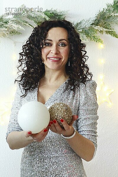 Porträt einer fröhlichen  positiven Frau  die ein glitzerndes Festtagskleid mit festlicher Frisur und Make up trägt und Weihnachtsbaumkugeln mit Ornamenten hält  eine glückliche Frau in stilvoller Weihnachtskleidung  die in die Kamera lächelt