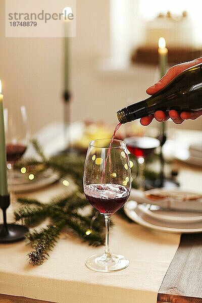 Brennende Kerzen und Gläser mit Rotwein in der Nähe von Tellern und Nadelbaumzweigen während der Weihnachtsfeier zu Hause. Crop anonyme männliche stehend am Tisch mit verschiedenen Gerichten und gießen Wein in Glas während Haus Party mit Freunden