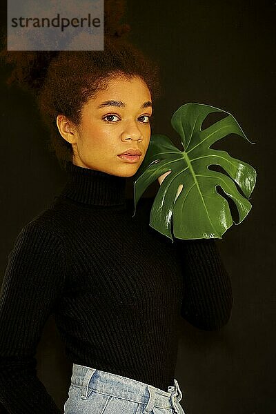 Natürliche Schönheit. Dunkles Studioporträt einer jungen afrikanisch amerikanischen Frau mit grünem Monsterablatt und Blick in die Kamera  nachdenkliche afrikanische Frau  die eine tropische Pflanze hält  isoliert über einer schwarzen Wand