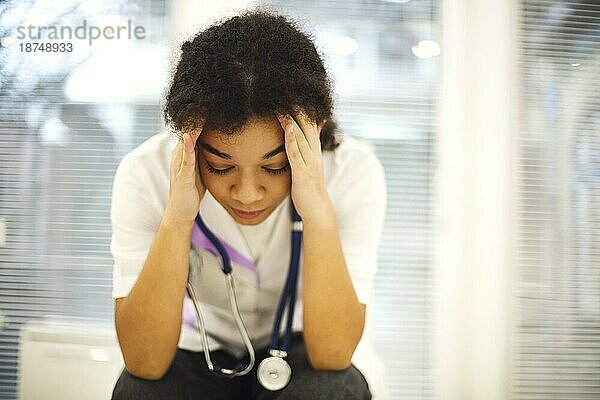 Traurige  deprimierte junge afroamerikanische Krankenschwester mit frustriertem Gesichtsausdruck  die sich von der Arbeitsbelastung in der Klinik überfordert fühlt  ethnische Ärztin  die sich nach einer anstrengenden Schicht müde fühlt