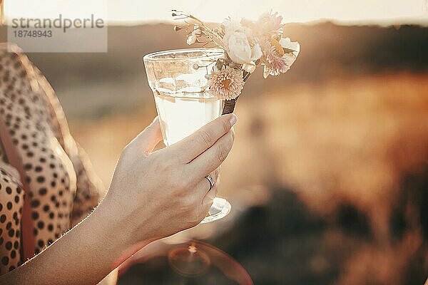 Cropped Schuss der Frau hält hohe Glas frisches Wasser mit kleinen Strauß von Wildblumen geschmückt  während in sonnigen Wiese bei Sonnenuntergang  ruhig und entspannend Natur Umgebung in verschwommenen Hintergrund stehen