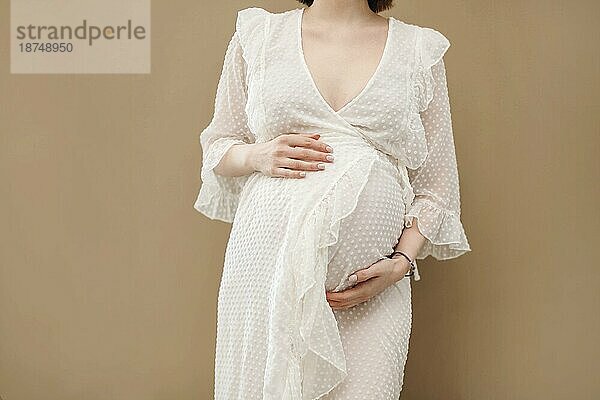 Attraktive schwangere Frau im weißen Kleid auf beigem Hintergrund. Mutterschaft und Gesundheitswesen Konzept. Nahaufnahme