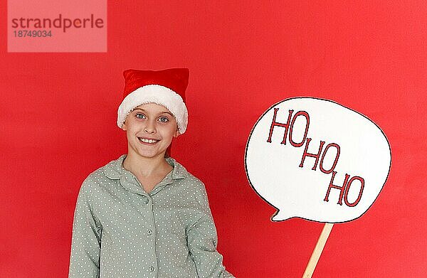 Frohe Feiertage. Nettes lächelndes kleines Mädchen in Santa Hut trägt Pyjama hält Board mit ho ho ho Text  Kind gratulieren mit Weihnachten und Neujahr  stehend isoliert über roten Hintergrund